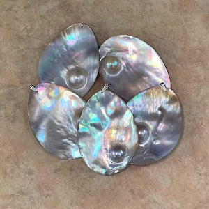 May include: Five iridescent, oval pendants with a pearlescent sheen. Each pendant has a small, round, pearl-like embellishment and a silver-coloured bail. The pendants are arranged on a neutral-toned surface.