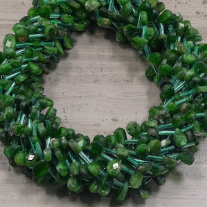 May include: A strand of green gemstone beads, likely emerald or peridot, with a faceted, teardrop shape. The beads are strung on a wire or thread and are arranged in a circular pattern.