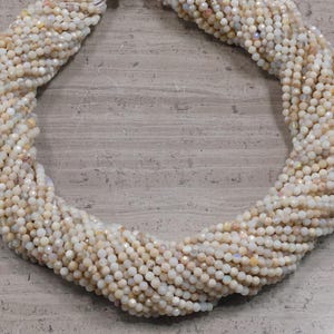 Puede incluir: Un collar de pequeñas cuentas redondas de color blanquecino con un brillo iridiscente sutil. Las cuentas están fuertemente ensartadas, formando una forma retorcida similar a una cuerda. Las cuentas tienen una superficie facetada.