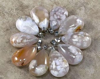 May include: A collection of ten teardrop-shaped pendants. The pendants are made of polished agate, displaying a mix of white, tan, and peach colors. Each pendant has a silver-colored bail for attaching to a chain. The pendants are arranged in a circular pattern.