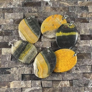 May include: Six oval-shaped pendants made of yellow, black, and gray bumblebee jasper. Each pendant has a silver bail for hanging.