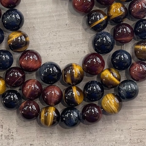May include: A strand of round, polished gemstone beads in shades of brown, gold, red, and blue. The beads are arranged in a circular pattern, creating a decorative and eye-catching display.