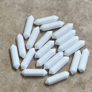 Könnte beinhalten: Eine Ansammlung von zwanzig kleinen, glatten, weißen, zylindrischen Perlen mit zwei Spitzen. Jede Perle hat an beiden Enden eine Spitze, was eine gleichmäßige, längliche Form erzeugt. Die Perlen sind in einer losen Gruppe angeordnet.