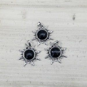 May include: Three silver sun pendant charms with black gemstone centers. The pendants are on a light gray background.