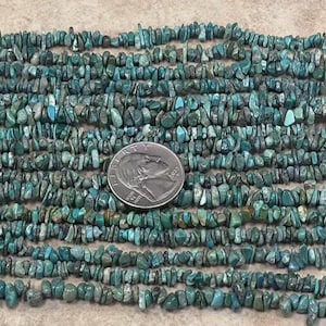 Op de afbeelding: Close-up van turquoise chip kralen, in verschillende tinten blauw en groen, gerangschikt in rijen. Een Amerikaanse kwart dollar munt is geplaatst op de kralen ter referentie. De kralen hebben een ruwe, onregelmatige vorm.