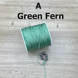 May include: A spool of green waxed cord with the text "A Green Fern" above it. A small string of dark brown square beads is strung on a length of cord.