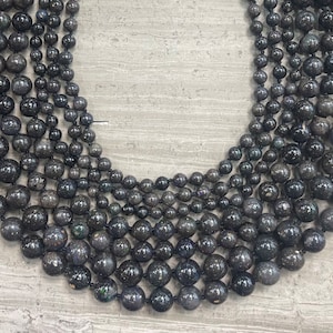 May include: A close-up of a strand of black gemstone beads. The beads are round and have a speckled appearance. The beads are strung on a black cord.