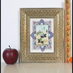 May include: An ornate framed miniature painting with a detailed gold and floral pattern. The painting depicts figures on horseback in a landscape scene. A red apple sits to the left of the frame. A ruler is next to the frame.