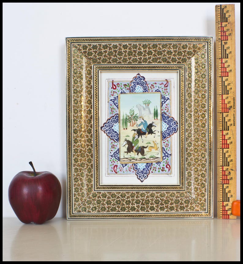 May include: An ornate framed miniature painting with a detailed gold and floral pattern. The painting depicts figures on horseback in a landscape scene. A red apple sits to the left of the frame. A ruler is next to the frame.