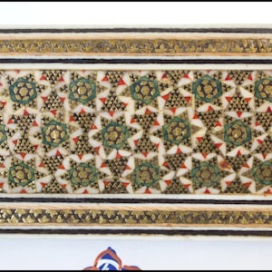 May include: Close-up of a decorative panel with an intricate geometric pattern. The design features gold, green, and red accents on a cream-colored background, framed by gold and black borders. The pattern is symmetrical and detailed.