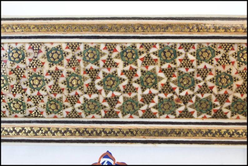 May include: Close-up of a decorative panel with an intricate geometric pattern. The design features gold, green, and red accents on a cream-colored background, framed by gold and black borders. The pattern is symmetrical and detailed.