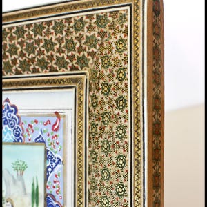 May include: Close-up of a decorative frame with intricate mosaic and gold detailing. The frame features a detailed pattern of green, red, and gold geometric shapes. The artwork within the frame depicts a scene with figures on horseback and landscape elements.