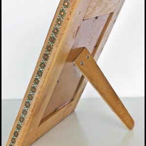 May include: Wooden picture frame with a decorative border featuring a repeating geometric pattern in green, orange, and brown. The frame has a built-in easel back for display. The frame is made of light-colored wood.