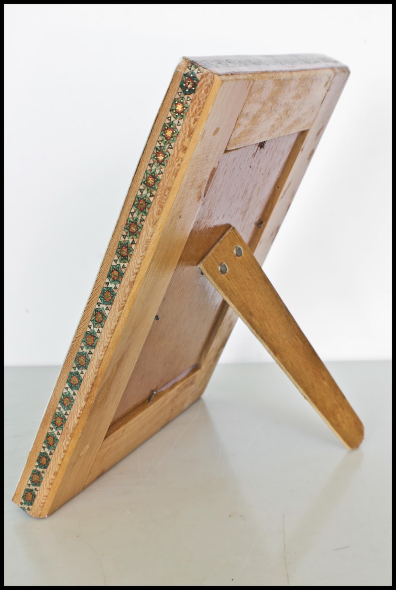 May include: Wooden picture frame with a decorative border featuring a repeating geometric pattern in green, orange, and brown. The frame has a built-in easel back for display. The frame is made of light-colored wood.