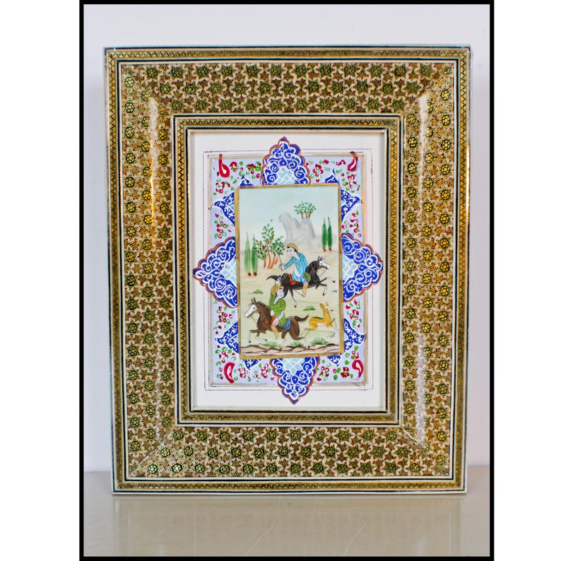 May include: An ornate framed artwork featuring a miniature painting of figures on horseback. The frame has a gold and green floral pattern, with a blue and white decorative border around the painting. The painting depicts a landscape scene.