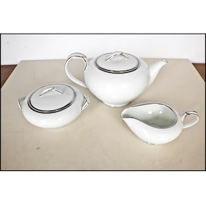 May include: A white porcelain tea set with silver trim. The set includes a teapot with a lid, a sugar bowl with a lid, and a creamer. The pieces are arranged on a neutral surface.