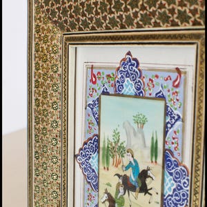 May include: An ornate framed artwork featuring a miniature painting of figures on horseback in a landscape. The frame has intricate gold and green floral patterns, with a blue and white decorative border.