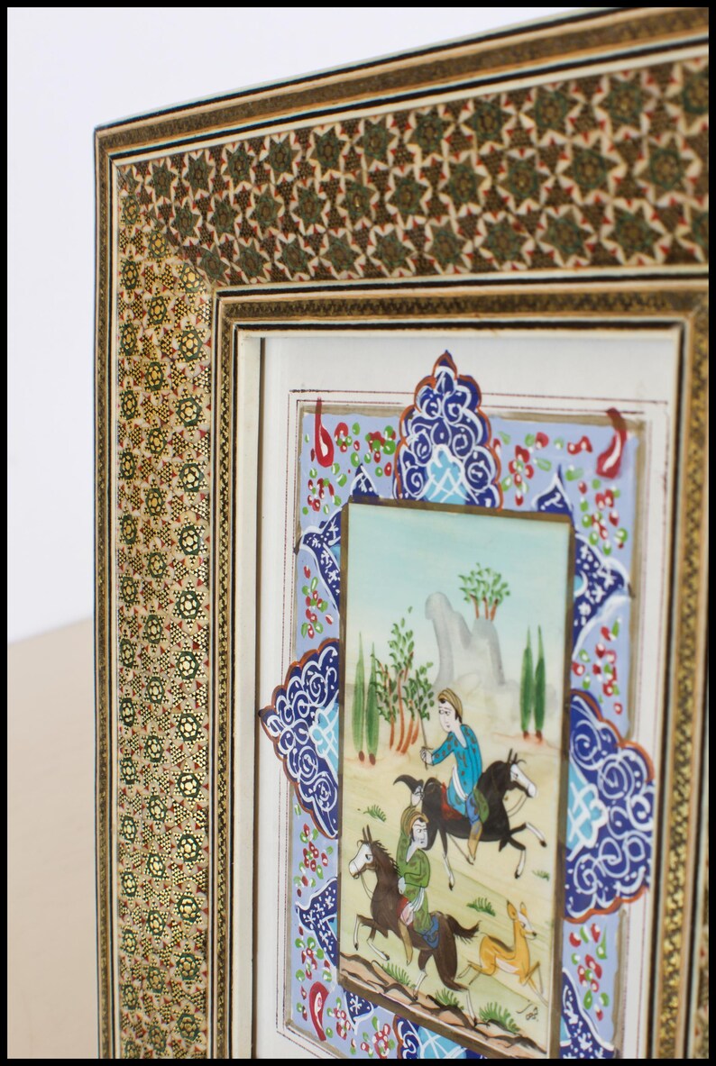 May include: An ornate framed artwork featuring a miniature painting of figures on horseback in a landscape. The frame has intricate gold and green floral patterns, with a blue and white decorative border.