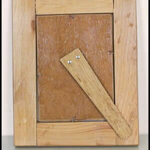 May include: Back view of a square wooden picture frame. The frame is made of light-colored wood with a natural finish. A wooden easel prop is attached to the back. The frame is designed to hold a photograph or artwork.