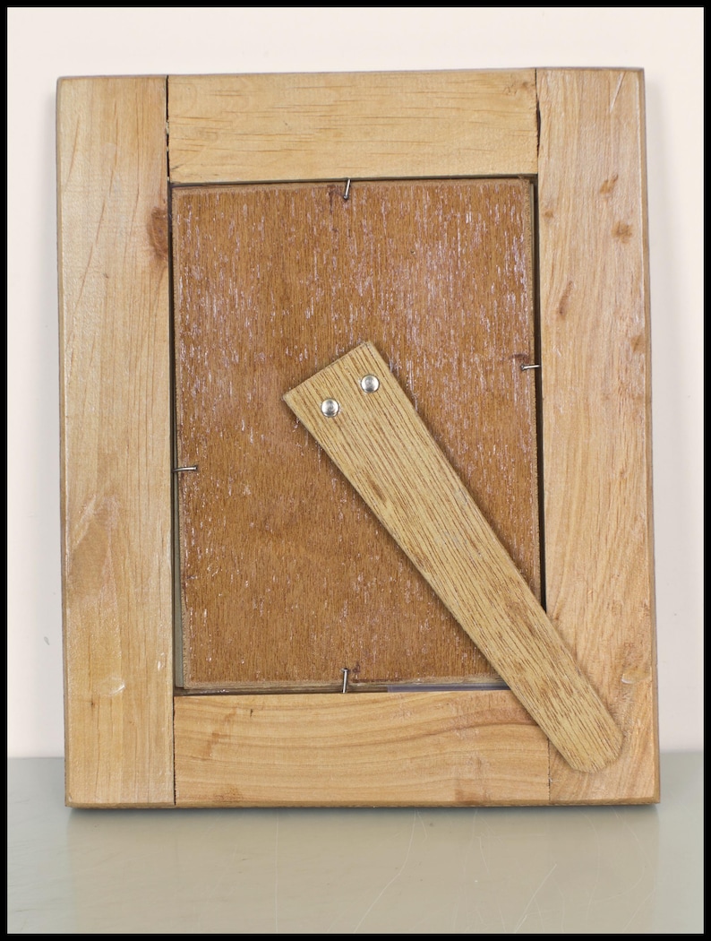 May include: Back view of a square wooden picture frame. The frame is made of light-colored wood with a natural finish. A wooden easel prop is attached to the back. The frame is designed to hold a photograph or artwork.