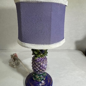 Puede incluir: Una lámpara de mesa decorativa con una pantalla morada y una base en forma de piña. La base está pintada en tonos de morado, verde y dorado. La pantalla es de color morado claro con un borde blanco. La lámpara tiene un cable de alimentación.