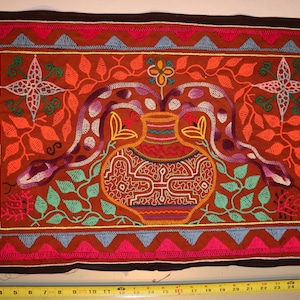 Może przedstawiać: Haftowany materiał z centralnym wzorem wazonu, otoczony przez dwa węże. Dzieło charakteryzuje się żywą paletą kolorów pomarańczowego, czerwonego, fioletowego i zielonego, z brązową obwódką. Element prezentuje skomplikowane szwy i geometryczny wzór obramowania.