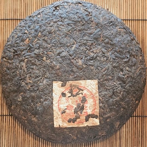 Puede incluir: Una torta de té redonda y marrón oscuro con una superficie texturizada. Una pequeña etiqueta rectangular con texto rojo y negro está adherida a la torta. La torta de té está sobre un fondo de estera de bambú.