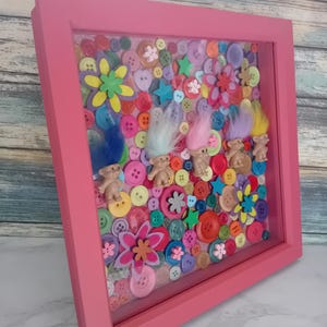 May include: A pink framed shadow box filled with colourful buttons, star and flower shapes, and small troll dolls with colourful hair. The buttons are in various sizes and colours, creating a vibrant and playful design.