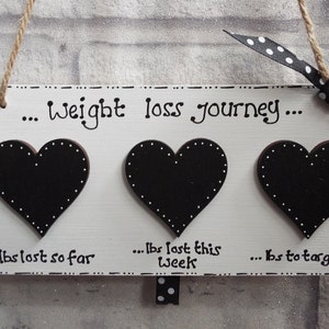 May include: White wooden sign with three black heart-shaped chalkboard pieces. The sign reads "... weight loss journey..." and has the text "... lbs lost so far", "... lbs lost this week", and "... lbs to target" written below the hearts.