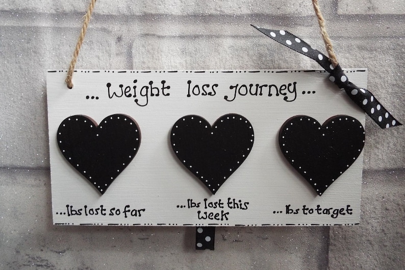 May include: White wooden sign with three black heart-shaped chalkboard pieces. The sign reads "... weight loss journey..." and has the text "... lbs lost so far", "... lbs lost this week", and "... lbs to target" written below the hearts.