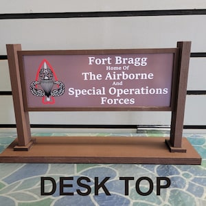 May include: A brown wooden sign with the text "Fort Bragg Home of The Airborne And Special Operations Forces" and a white and gray military insignia. The sign is on a brown wooden base with the text "DESK TOP" in black letters.