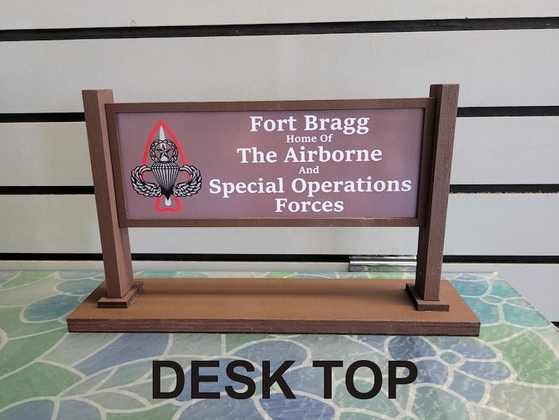 May include: A brown wooden sign with the text "Fort Bragg Home of The Airborne And Special Operations Forces" and a white and gray military insignia. The sign is on a brown wooden base with the text "DESK TOP" in black letters.