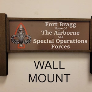May include: A brown wooden sign with a white background and the text "Fort Bragg Home of The Airborne And Special Operations Forces". The sign also features a black and white image of a parachute and a star. The sign is labeled "WALL MOUNT".