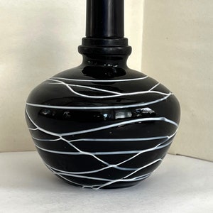 Może przedstawiać: Czarna szklana butelka o zaokrąglonym korpusie i cylindrycznym korku. Butelka jest ozdobiona białymi, falistymi liniami, które owijają się wokół jej powierzchni. Projekt nadaje butelce nowoczesny, artystyczny wygląd. Butelka jest prawdopodobnie używana do perfum lub olejków eterycznych.