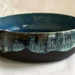 May include: A round ceramic bowl with a matte black exterior base and a glossy interior glaze that transitions from deep blue to a textured, dripped turquoise and white pattern along the upper rim and outer edge of the vessel.