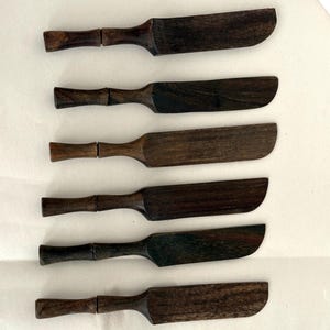 May include: Six dark brown wooden butter knives with rounded blades and textured handles are arranged on a white surface. The knives have a rustic, handcrafted appearance, suggesting they are part of a kitchen or dining set.