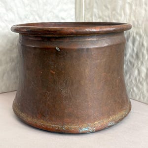 May include: A copper-colored, cylindrical container with a rounded rim. The vessel has a slightly flared base and shows signs of age and use, with variations in color and texture. The metal has a warm, reddish-brown hue with some areas of patina.