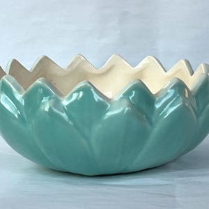 May include: A ceramic bowl with a teal exterior and a cream-colored interior. The bowl has a unique petal-like design around the rim. The glossy finish enhances the colors. This decorative bowl is suitable for various uses.