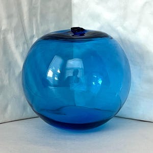 Puede incluir: Un objeto decorativo redondo de vidrio azul con una parte superior azul oscuro. La esfera de vidrio refleja el entorno circundante. El objeto está sobre una superficie blanca.