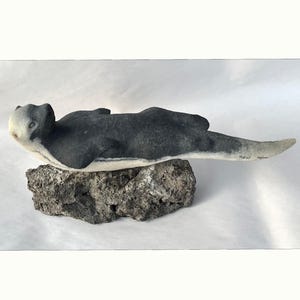 Peut inclure: Sculpture artisanale d'une loutre de mer, allongée sur un rocher gris texturé. La loutre a un corps gris et blanc avec un visage détaillé. La sculpture est une œuvre d'art unique.