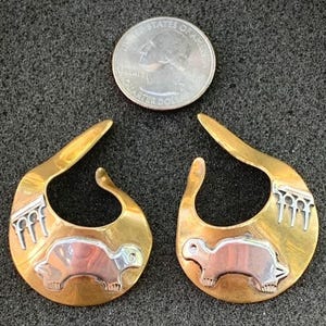 May include: A pair of gold hoop earrings with a silver turtle design and a silver geometric pattern on the side. The earrings are about the size of a UK 20 pence coin.