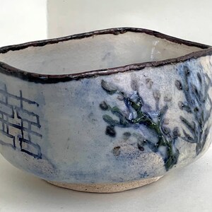 May include: A hand-formed ceramic bowl with an irregular, organic shape and a dark, textured rim. The exterior features a light blue and white glaze with a hand-painted blue double happiness symbol and a dark blue branch motif with small buds.