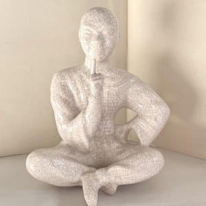 May include: A ceramic figurine of a person in a seated meditation pose, with a finger raised to the lips. The figure is off-white with a cracked, aged appearance. The figurine is a decorative item.