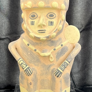 Puede incluir: Una figura de cerámica de color beige y gris de una figura estilizada con una cabeza grande y brazos a los lados. La figura tiene marcas negras en la cara y los brazos, y un collar con puntos negros. La figura está sobre un soporte transparente.