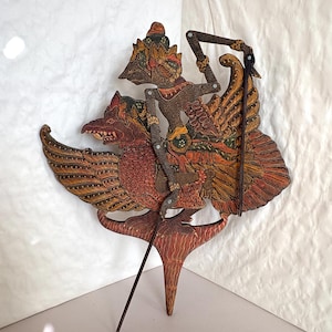 May include: A detailed, hand-carved Indonesian shadow puppet depicting a figure riding a mythical bird. The puppet features intricate patterns in shades of brown, red, and gold. The bird has large, detailed wings, and the rider is adorned with ornate details.
