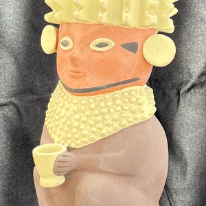 Puede incluir: Una figura de cerámica de una figura sentada con un cuerpo marrón claro, sosteniendo una pequeña taza. La figura tiene una cara bronceada, un cuello amarillo y un tocado amarillo. La figura se exhibe sobre un soporte rectangular transparente.
