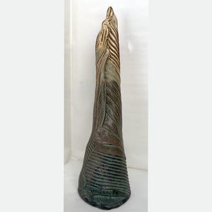May include: A tall, textured ceramic sculpture with a vertical, elongated form. The sculpture features a blend of brown, green, and beige tones, with linear and textured patterns. The base is wider, tapering upwards to a pointed top.