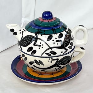 Może przedstawiać: Ceramiczny zestaw do herbaty z pasującym spodkiem. Czajniczek jest biały z czarnymi abstrakcyjnymi wzorami i niebieską, zieloną i różową pokrywką. Spodek ma wzór w paski w różnych kolorach. Zestaw przeznaczony jest do serwowania herbaty.