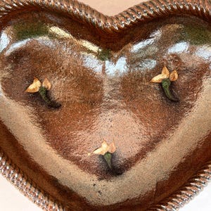 May include: Heart-shaped ceramic bowl with a glossy brown interior and a textured, rope-like border. The bowl features three small, decorative floral accents. A unique piece for serving or display.