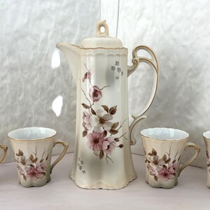 Op de afbeelding: Een porseleinen koffieservies bestaande uit een hoge koffiepot met deksel en vier bijpassende kopjes. Elk stuk heeft een crèmekleurige basis met een geschulpte rand, goudkleurige accenten en een delicaat bloemmotief van roze bloesems.